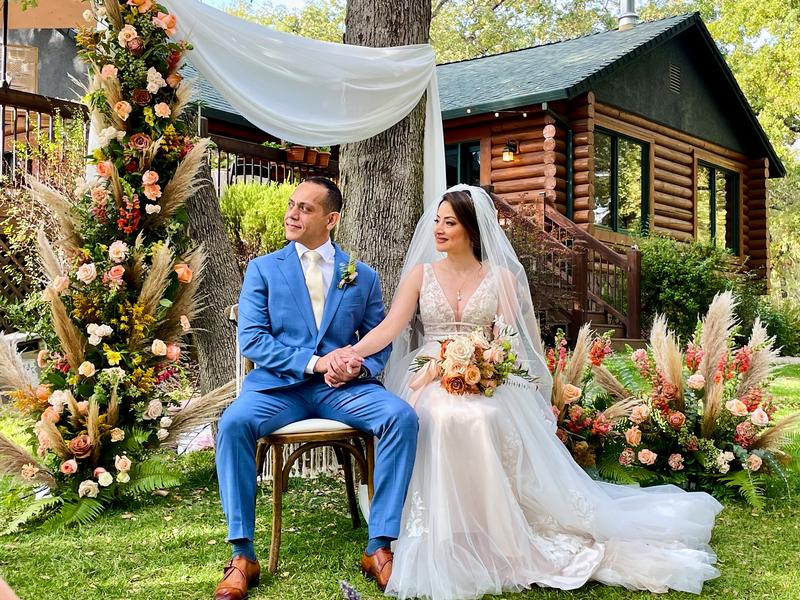Bride and groom seated hand in hand before a floral arch of roses and pampas grass