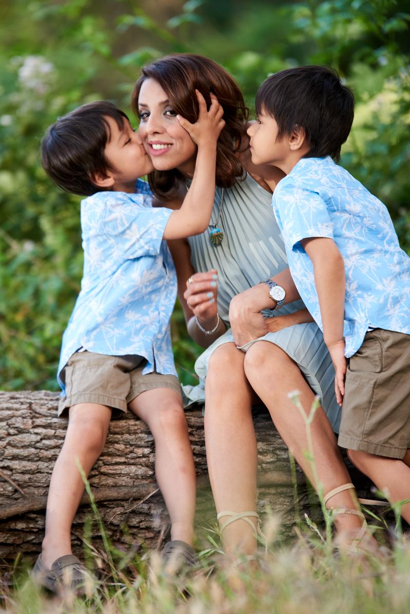 Two boys kissing their mother's cheek as she sits on a log